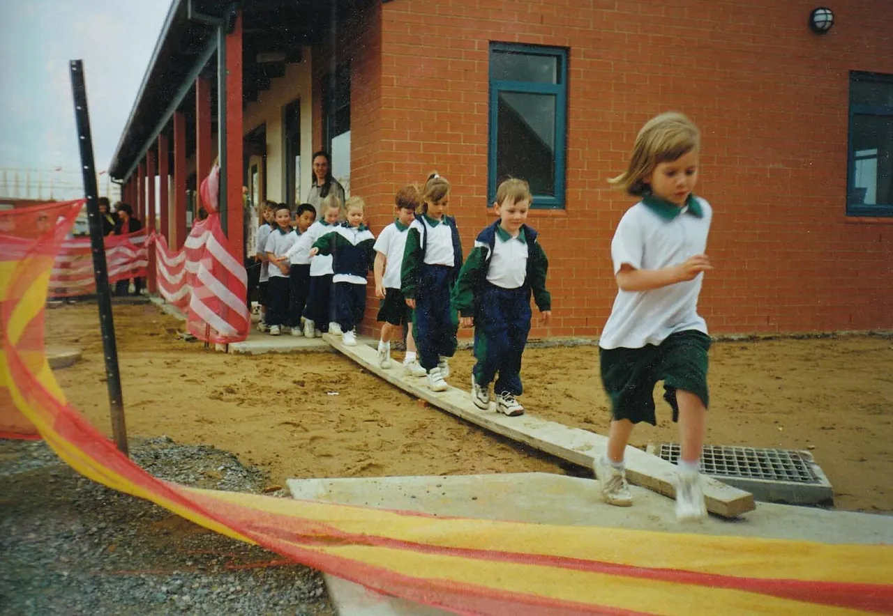 Kindergarten students running across a wooden plank over dirt to get between classrooms