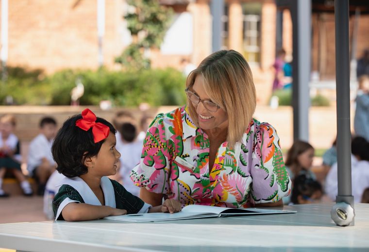 Primary school student reading a book with teacher outdoors.