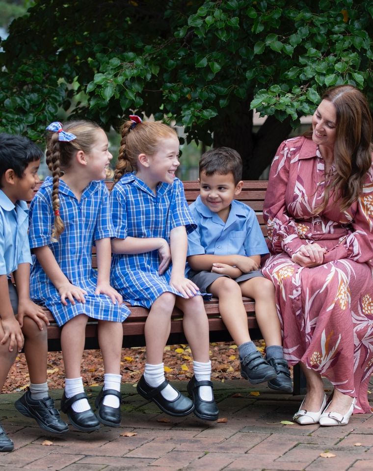 Primary school students sitting on bench outdoors with school principal.