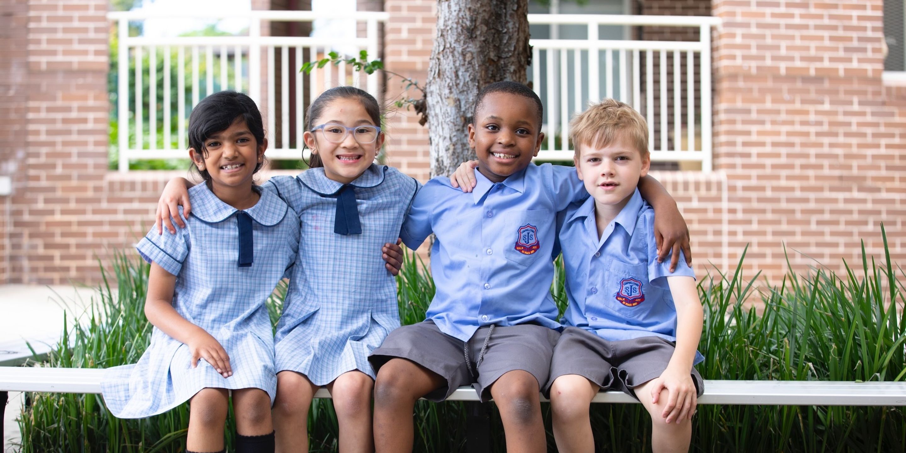 Primary school students sitting on bench outdoors with arms around each other.