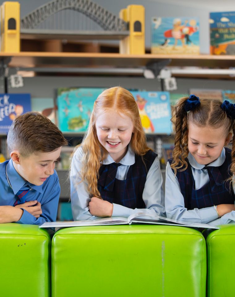 Three primary school students reading a book on green ottomans.