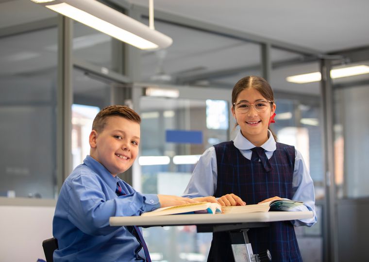 Two primary school students reading a book on desk and smiling at camera.