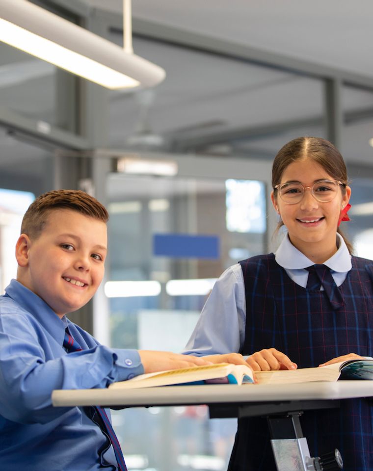 Two primary school students reading a book on desk and smiling at camera.