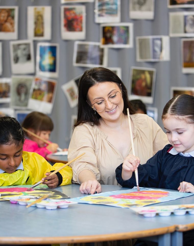 Two primary school students painting at desk with teacher.