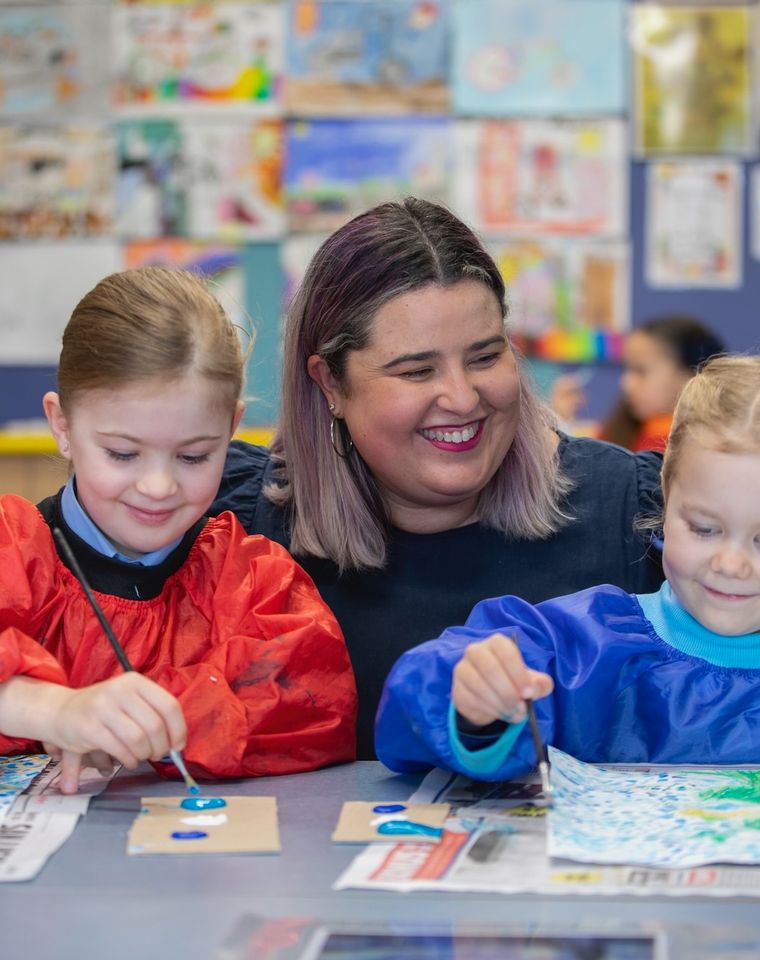 Two primary school students painting with teacher smiling between them.