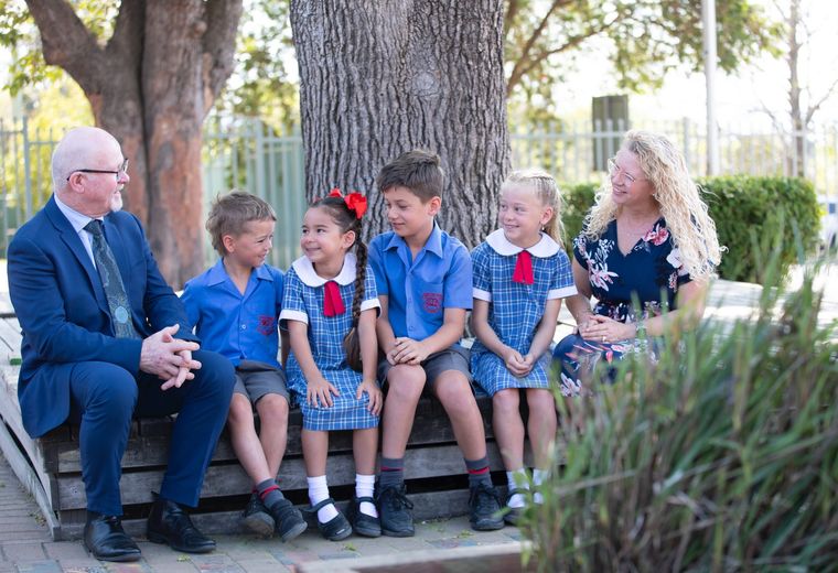 Primary school students sitting under tree outdoors with teacher and school principal.