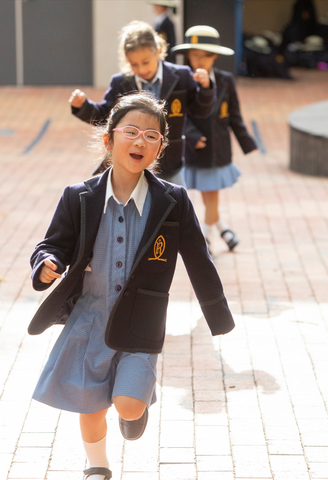 A Junior School student running and laughing in the playground at Ruyton Girls' School. She is wearing summer uniform.