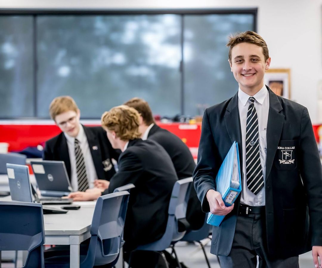 Student holding a binder, smiling at the camera with a group of students working together in the background