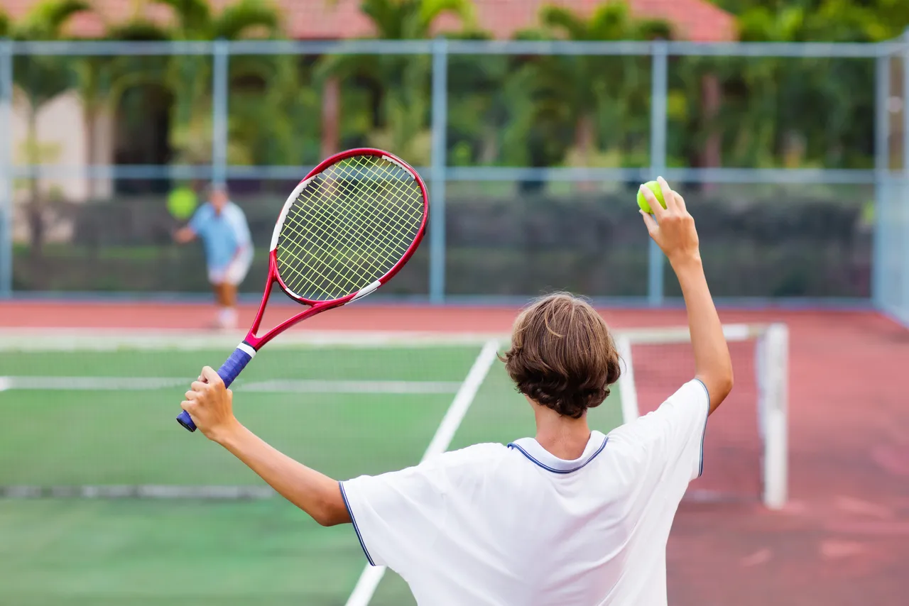 Boy playing tennis on outdoor court. Teenager with tennis racket and ball in sport club. Active exercise for kids. Summer activities for children. Training for young kid. Child learning to play.