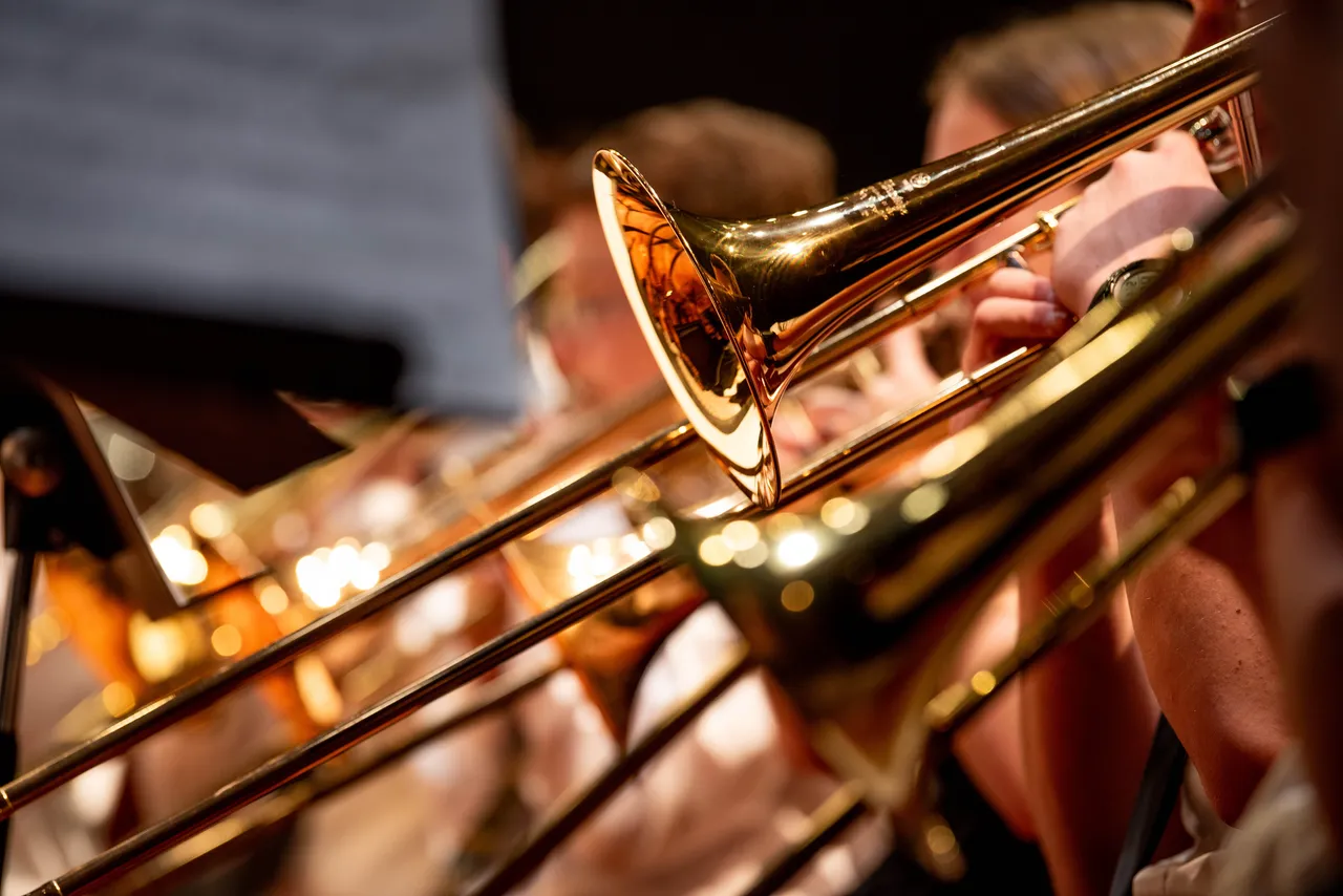 Close-up view of high school band students performing with brass instruments, including trumpets and trombones, during a school concert. The image captures the golden metallic shine of the instruments with warm stage lighting, showcasing youth musical education and ensemble performance. Perfect for illustrating music education, school programs, student achievement, and performing arts in educational settings.