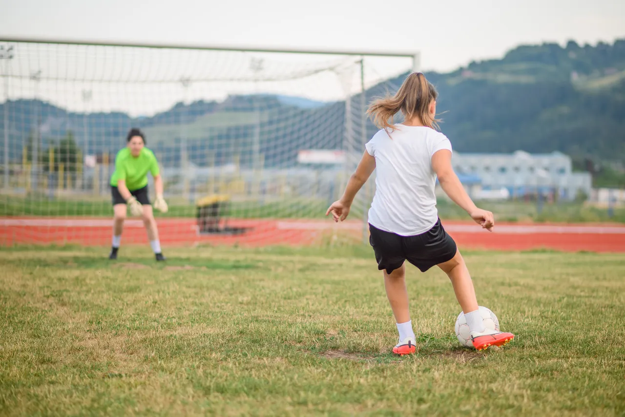 Teenage girl practicing soccer, kicking the ball towards the goal with the goalkeeper ready to defend