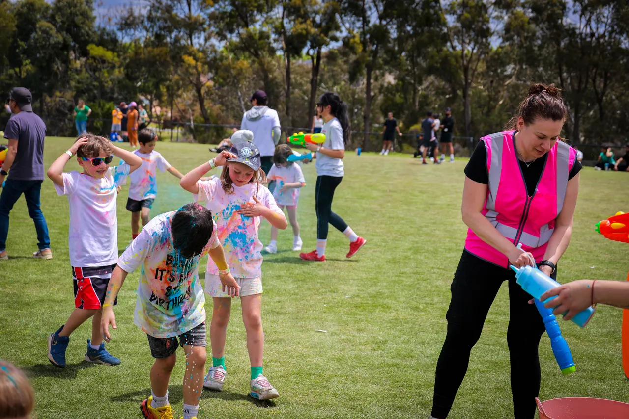Overnewton Community Engagment Program parent volunteer supporting bi annual Parents and Friends Association fundraising Colour Fun Run held at Canowindra Campus, Taylors Lakes, as primary school-aged children run past.