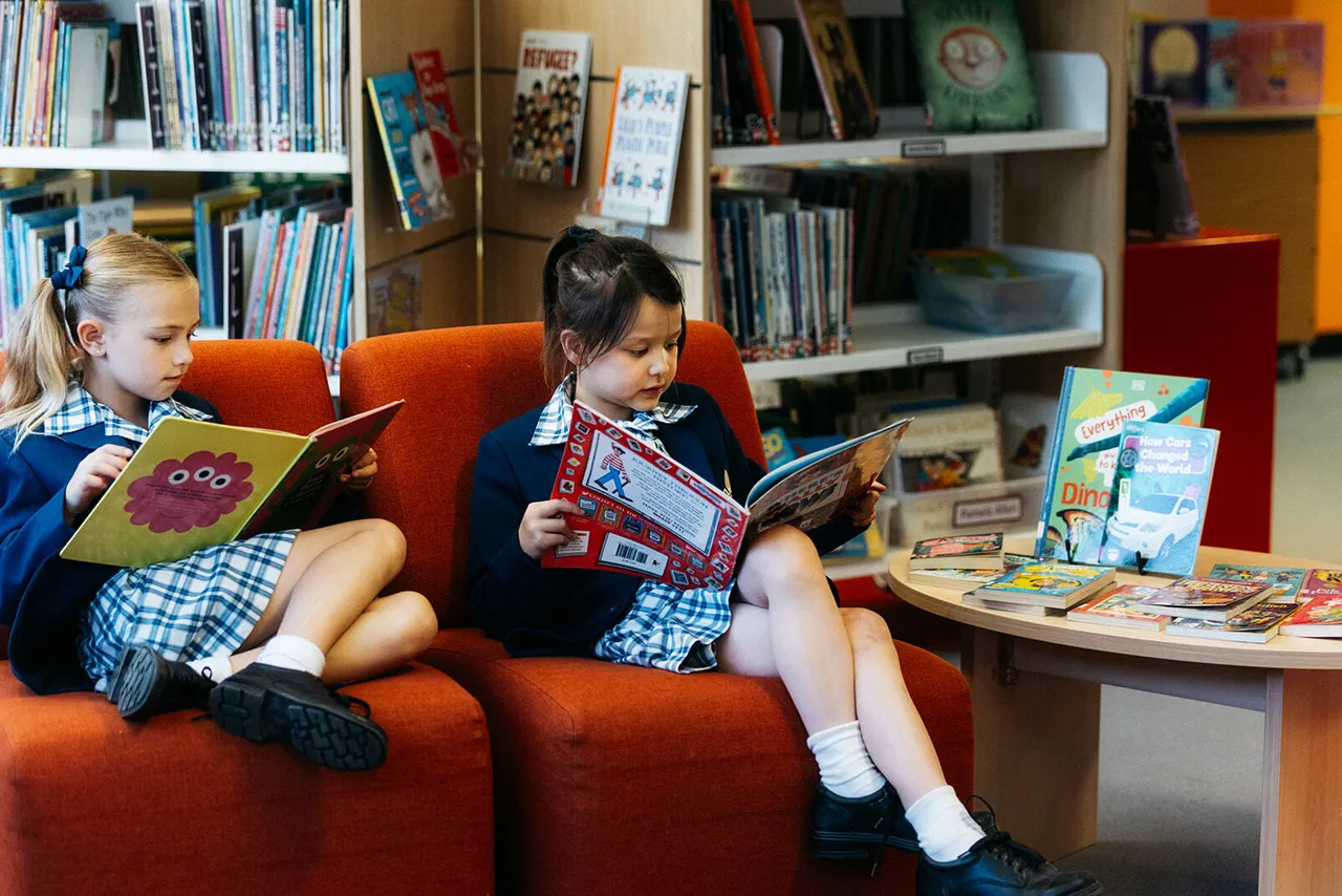 Two lower primary school aged girls reading in the Junior School library at Canowindra Campus, sitting on colourful red couches surrounded by books.