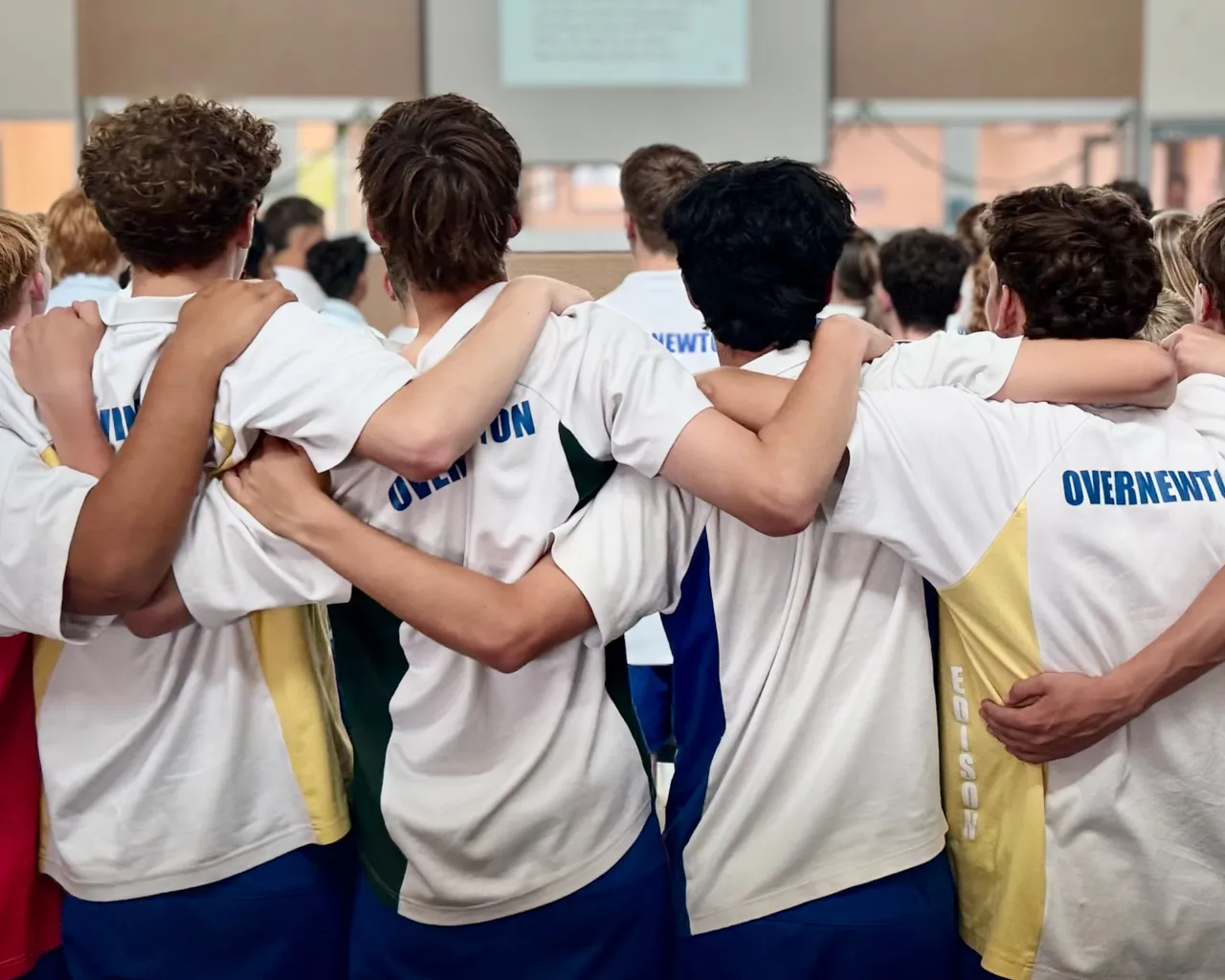 Action shot from behind of Year 9 secondary school aged students wearing the Overnewton Anglican Community College sports uniform, linked arm-in-arm in solidarity as they gather to celebrate the end of the school year.