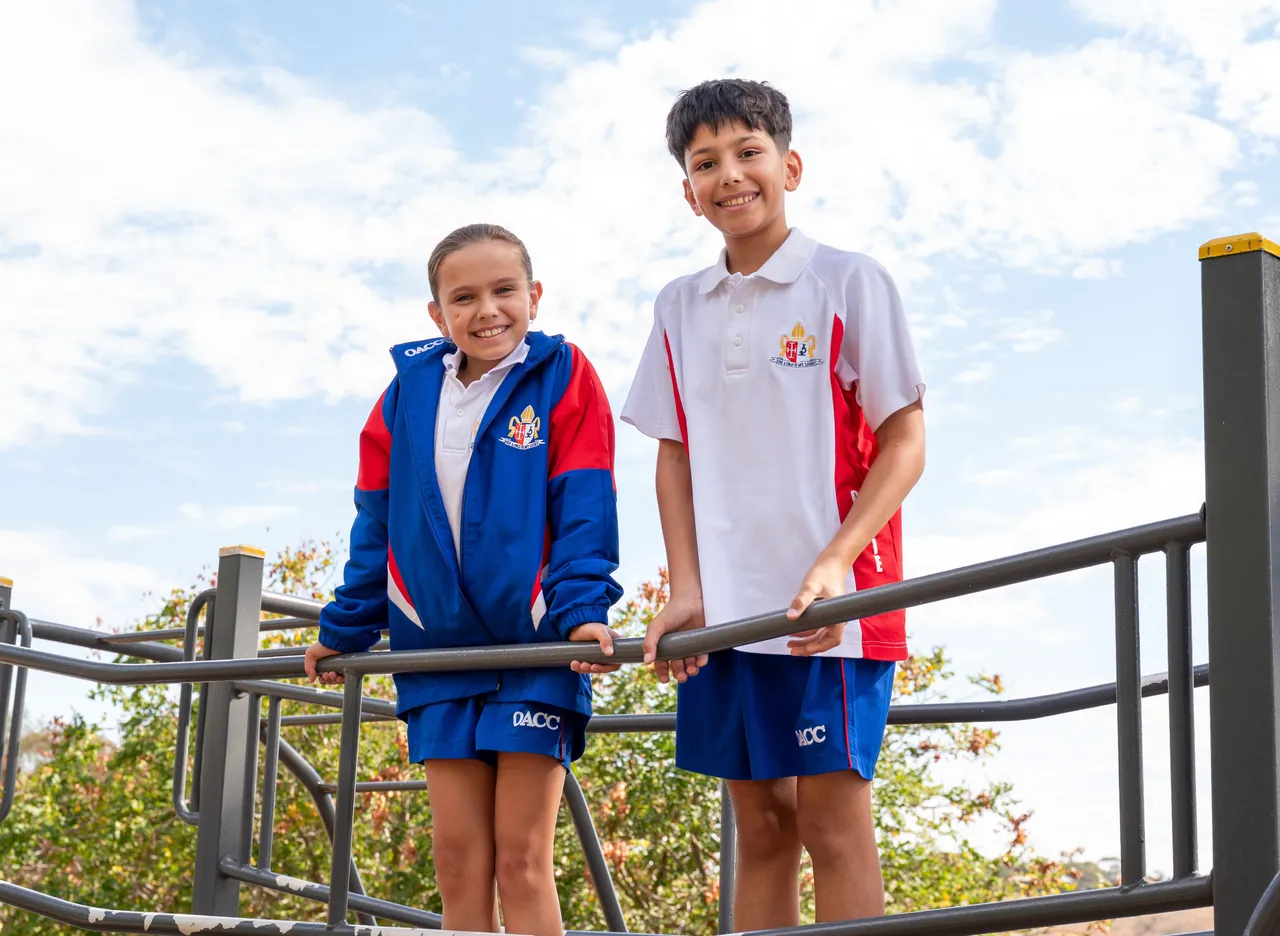 Two upper primary school aged students in sports uniform standing on a play bridge, surrounded by natural outdoor elements.