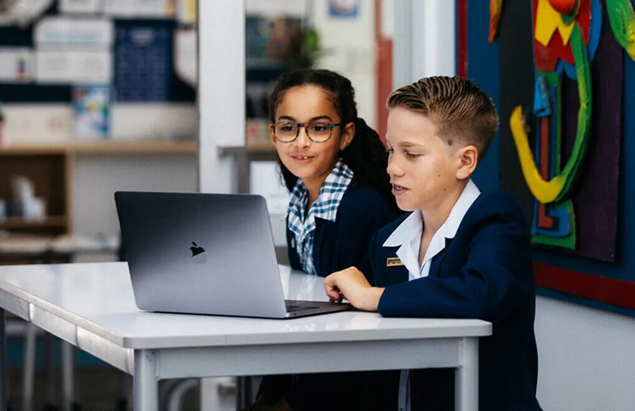 Two upper primary school aged students in an internal learning space at Yirramboi Campus, working on a MacBook in front of bright, colourful artwork.