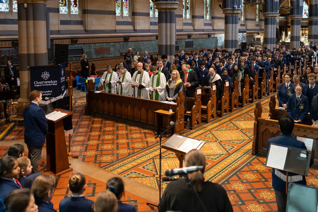 High angle shot from above the stage showing an upper secondary school aged student from Overnewton's Senior School standing at a lecturn while clergy in robes and members of the College Leadership Team watch from pews as part of the College's annual Founders' Day service.