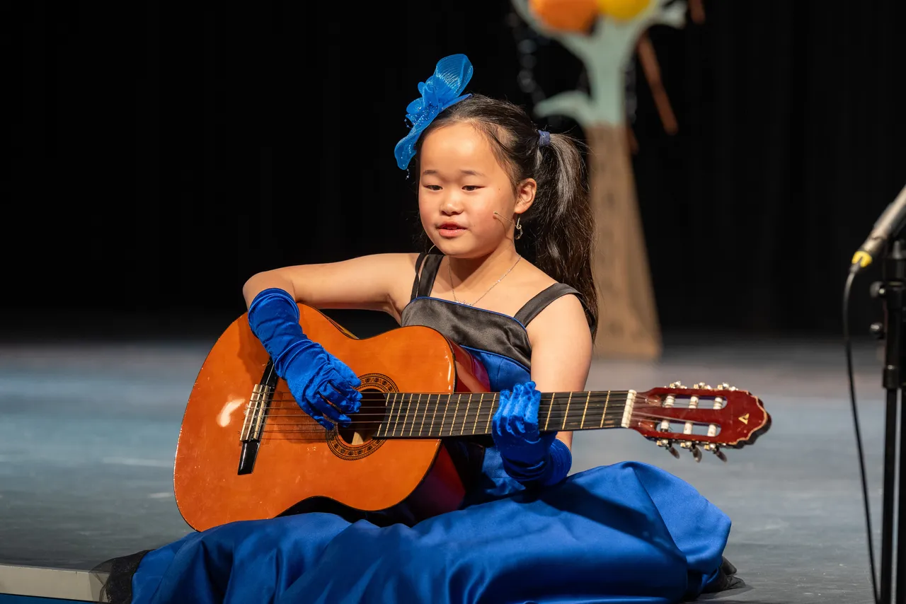 Upper primary school aged student wig captured holding a guitar and singing to the audience during the Middle School Year 5 & 6 Musical Season 1 production of Seussical KIDS.