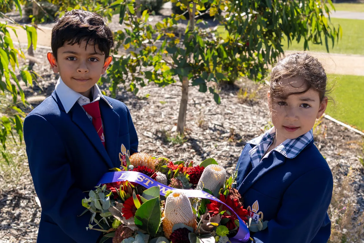 Middle primary school aged students from Overnewton's Junior School proudly hold a large commemorative wreath adorned with a "Lest We Forget" ribbon between them, which will be used as part of an ANZAC Day ceremony at the school.