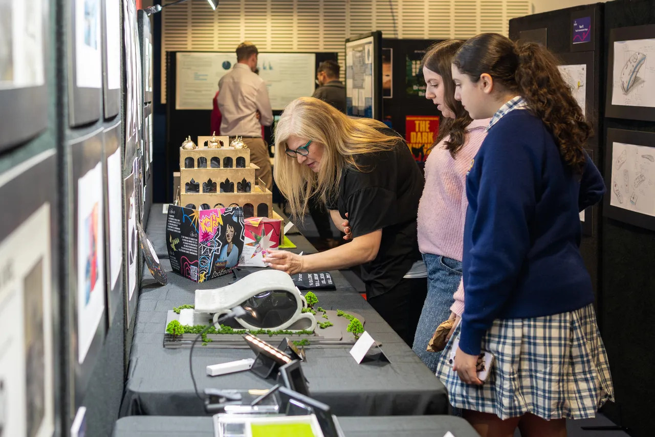 A guest at Overnewton's Annual Visual Arts & Technologies Exhibition viewing 3D models crafted Senior School Technologies students as two students look on.