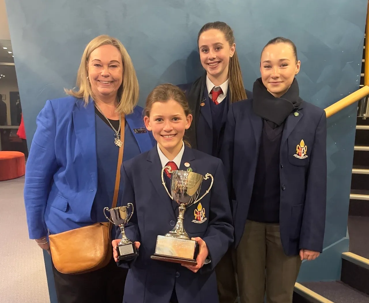 Three secondary school-aged students in uniform standing beside Principal Emily FitzSimons proudly holding two trophies for wins at the 2025 ACS Public Speaking Championships.