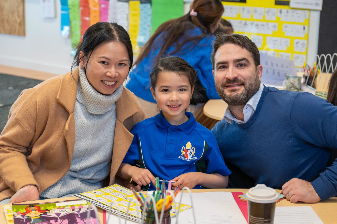 Lower primary school aged student proudly posing with her mother and father at her classroom desk during the Junior School Celebration of Learning event.