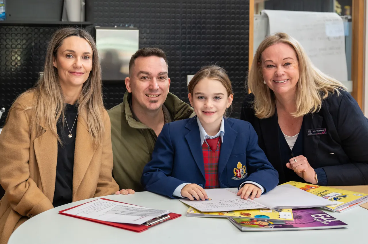 Lower primary school aged student proudly posing with her mother and father, plus College Principal Ms Emily FitzSimons, at her classroom desk during the Junior School Celebration of Learning event.