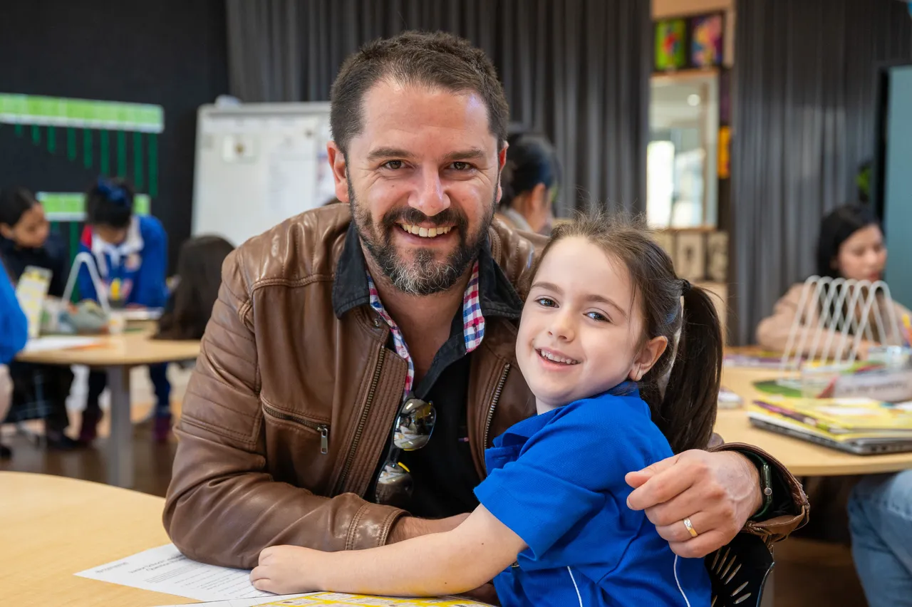 Lower primary school aged student proudly posing with her father at her classroom desk during the Junior School Celebration of Learning event.