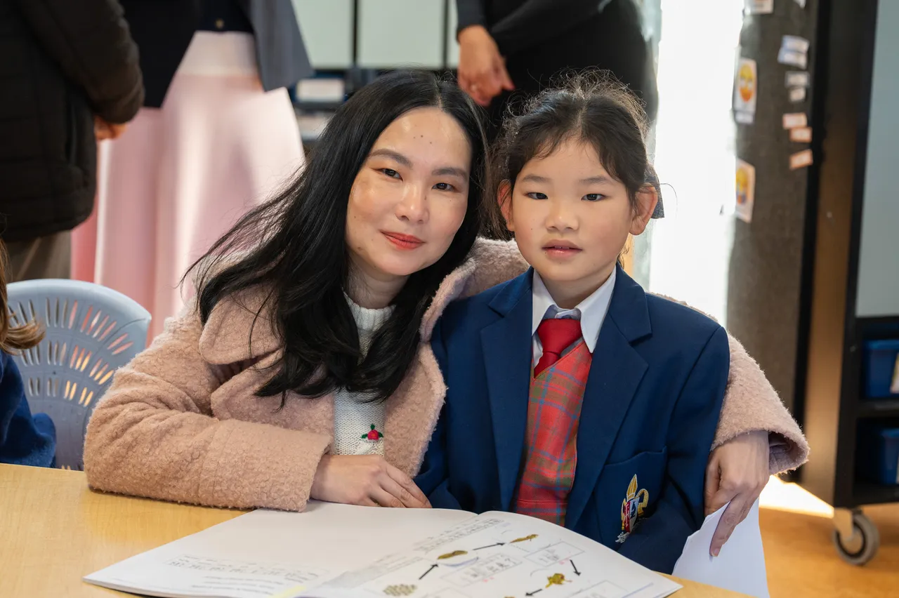 Lower primary school aged student proudly posing with her mother at her classroom desk during the Junior School Celebration of Learning event.