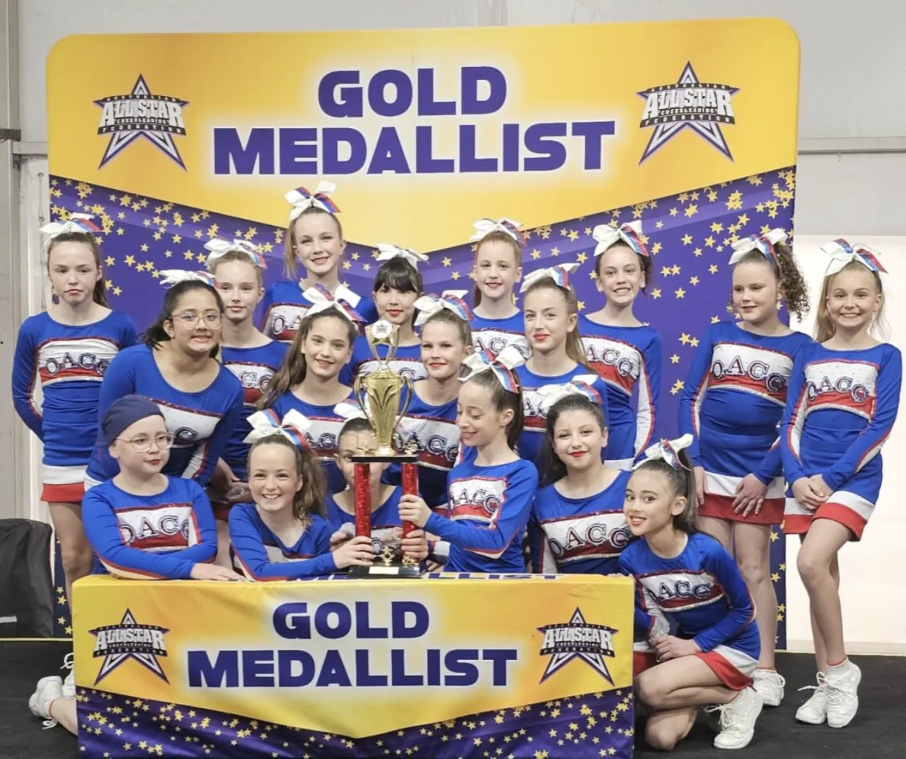 Upper primary school-aged students from the Overnewton Middle School Cheer team, Angels, celebrating in front of a Gold Medallist banner at the 2025 AASCF State Championships.
