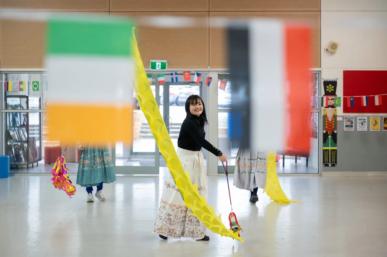 College Head of Languages, Sunny Huang, dressed in traditional Chinese attire performing a dragon dance with Year 9 secondary school aged students during Overnewton’s 2025 Languages and Culture Week, photographed through blurred flags in the foreground.