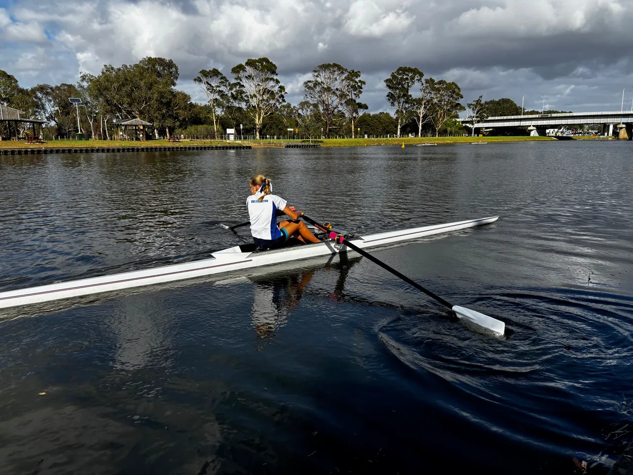 Image of senior secondary school aged student wearing her Overnewton sports shirt, in motion rowing on a wide river in a single skull craft.