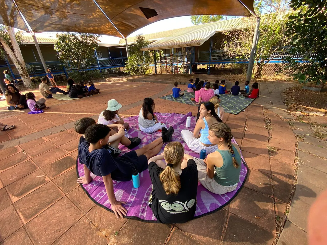 A group of Overnewton students and local students from Shepherdson College sitting on mats under a shade sail at Elcho Island in the Northern Territory during Overnewton’s 2025 Galiwin’ku educational travel program.
