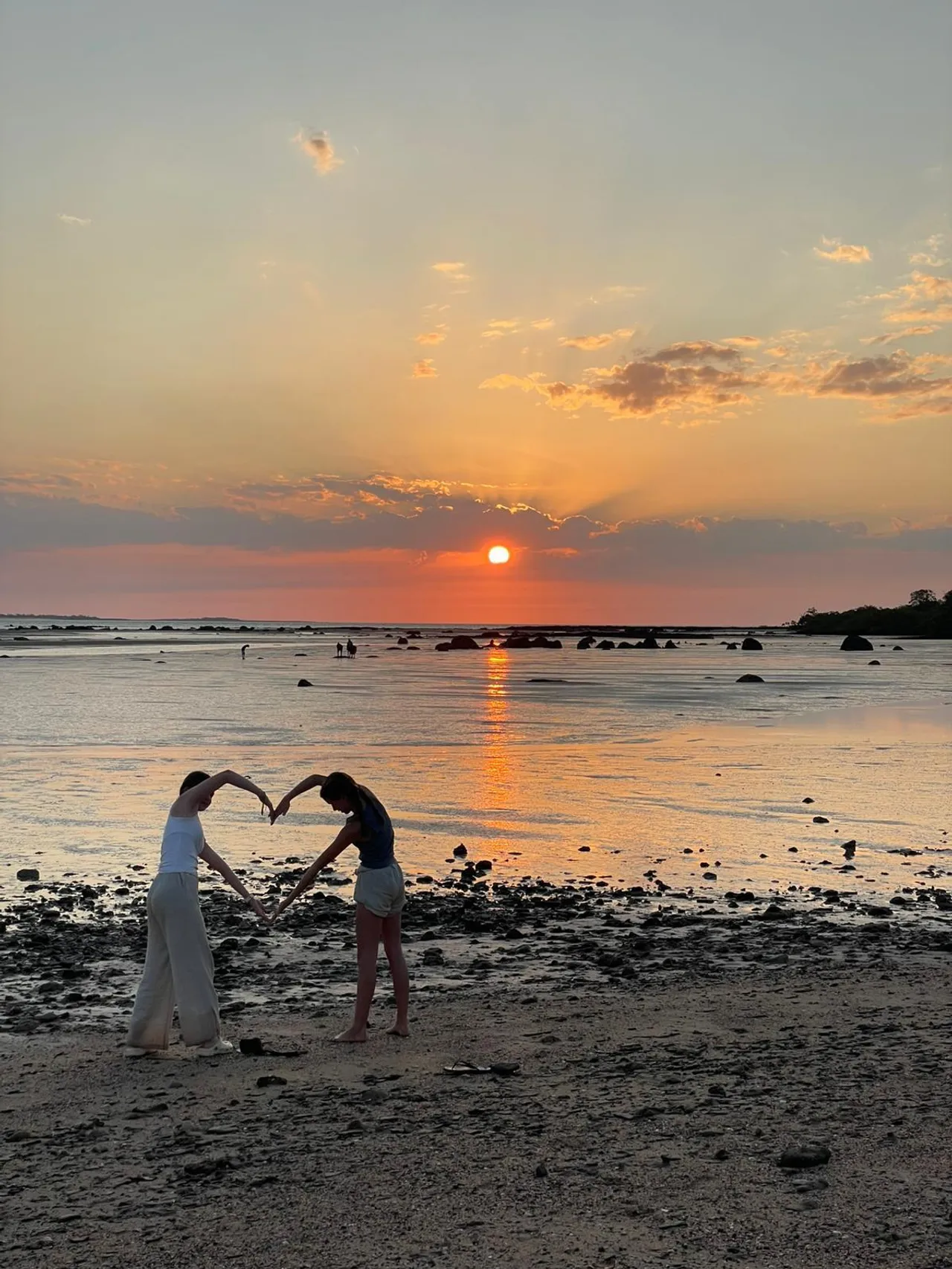 Two students joining their bodies at the hands to make a large heart shape as they stand at the water's edge on a sunset-lit beach at Elcho Island in the Northern Territory during Overnewton’s 2025 Galiwin’ku educational travel program.