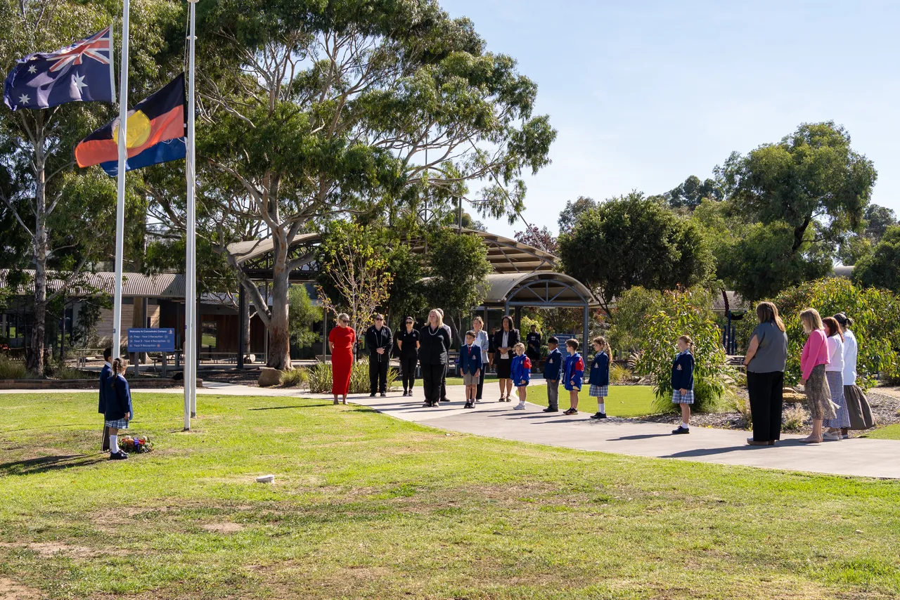 Middle primary school aged students from Overnewton's Junior School standing beside the flagpole with flags at half mast, while a group of observers watch on as part of the 2026 ANZAC Day commemoration at Canowindra Campus, Taylors Lakes.
