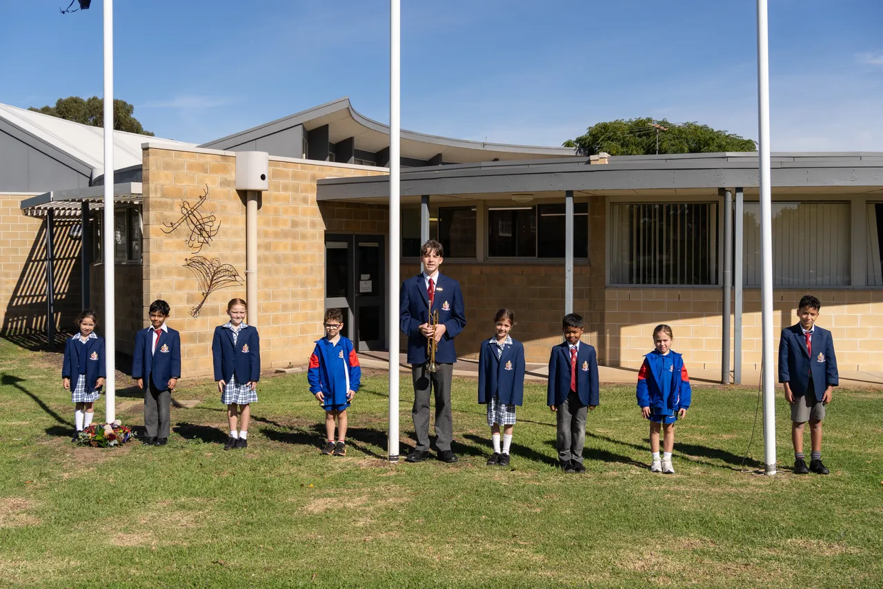 Middle primary school aged students from Overnewton's Junior School, and a Year 9 student bugle player, standing ceremoniously by the flagples at Canowindra Campus, Taylors Lakes, as part of the 2026 ANZAC Day commemoration.