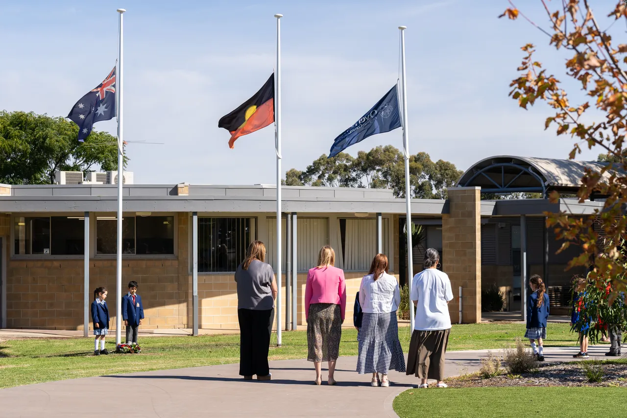 Middle primary school aged students from Overnewton's Junior School standing beside flagpoles with flags at half mast, while a group of observers watch on as part of the 2026 ANZAC Day commemoration at Canowindra Campus, Taylors Lakes.