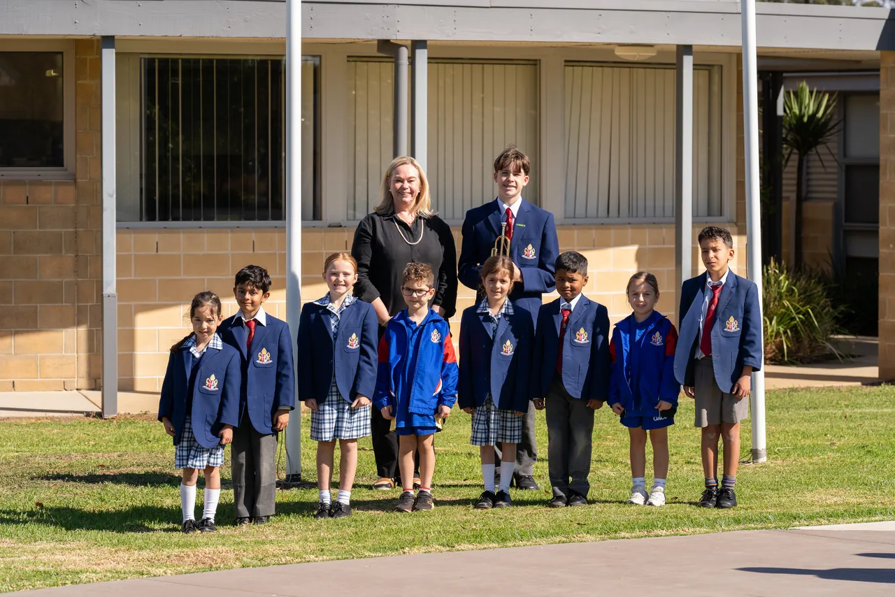 Middle primary school aged students from Overnewton's Junior School, a Year 9 student bugle player and Principal Emily FitzSimons, standing ceremoniously by the flagples at Canowindra Campus, Taylors Lakes, as part of the 2026 ANZAC Day commemoration.