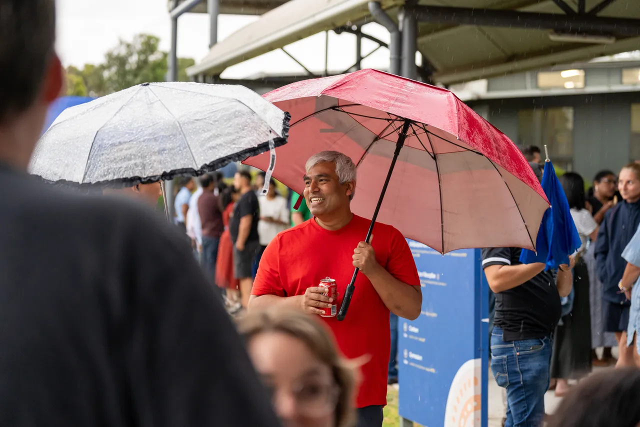 Member of the Overnewton community walking under a large red umbrella through the crowd at the sausage sizzle during the inaugural Canowindra Parent Information Evening.