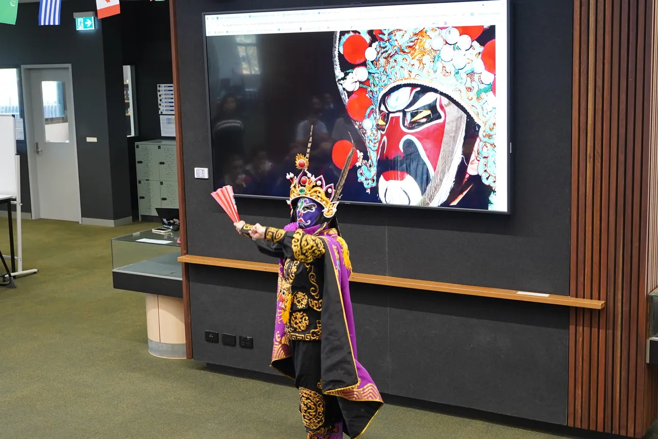 An expert face-changing Sichuan Opera artist in traditional dress performing extravagantly with a large fan for Middle School students at Yirramboi Campus during Overnewton's 2025 Languages and Culture Week.