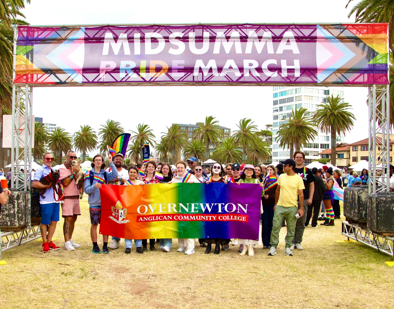 A group of upper secondary school aged students and staff representing Overnewton Anglican Community College at the 2026 Midsumma Pride March.  Group is assembled beneath the official event banner, with members of the group proudly holding a rainbow-coloured Overnewton school banner.