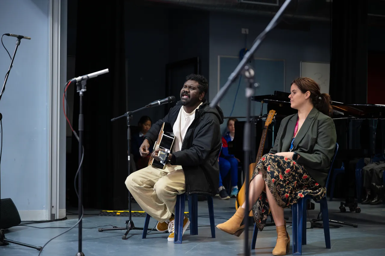 Highly regarded Indigenous musician, Yirrmal, playing a guitar and singing on the Sports Hall stage while College Head of Synergy, Sasha Koomen, watches on as part of the Overnewton Anglican Community College 2025 Yirramboi Campus Reconciliation Week Assembly.
