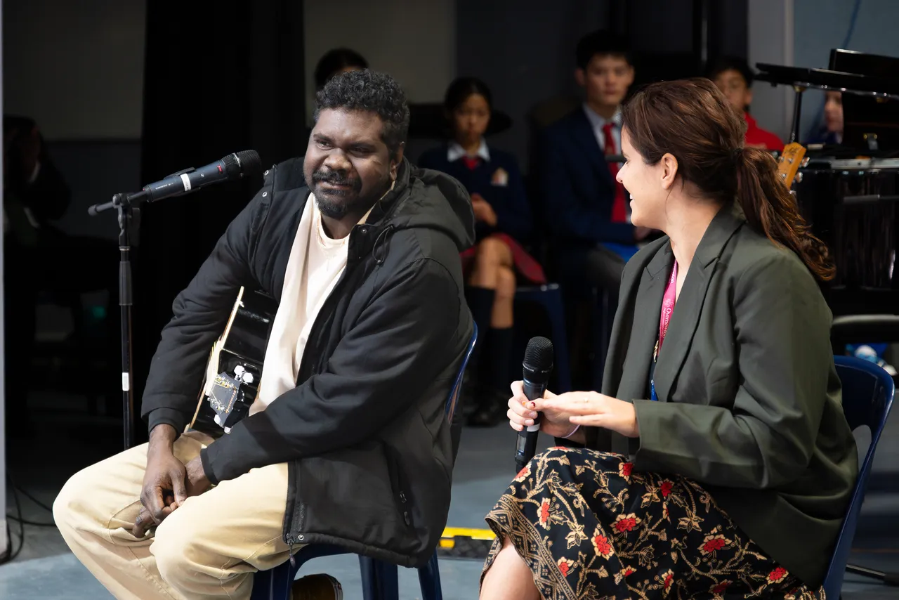 Highly regarded Indigenous musician, Yirrmal, holding a guitar on stage and engaged in conversation with College Head of Synergy, Sasha Koomen, as part of the Overnewton Anglican Community College 2025 Yirramboi Campus Reconciliation Week Assembly.
