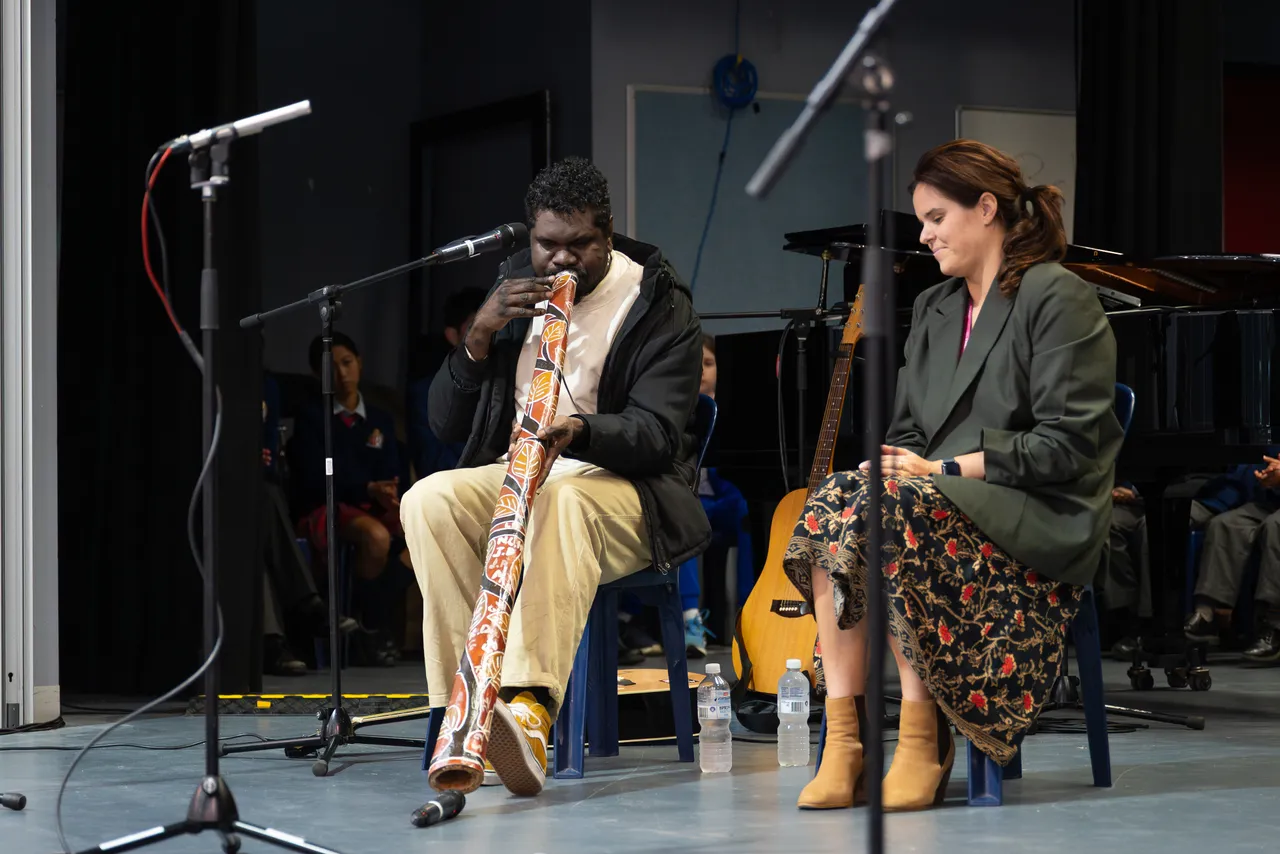 Highly regarded Indigenous musician, Yirrmal, playing a painted didgeridoo on stage while College Head of Synergy, Sasha Koomen, listens intently as part of the Overnewton Anglican Community College 2025 Yirramboi Campus Reconciliation Week Assembly.