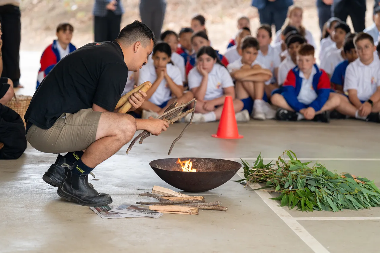 Proud member of the Wurundjeri people of the Kulin Nation holding a eucalyptus branch and kneeling beside a lit fire pit. Students can be seen sitting in the background watching as part of an annual Smoking Ceremony for Year 5 students new to the Yirramboi Campus.