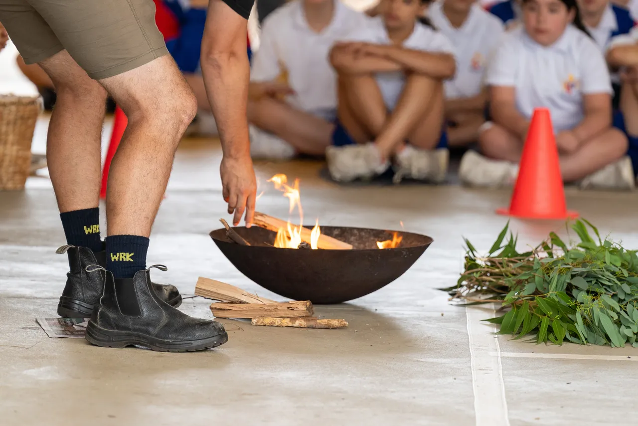 Thane, proud member of the Wurundjeri people of the Kulin Nation, tends to a lit fire pit. Students can be seen sitting in the background watching preparations to commence an annual Smoking Ceremony for Year 5 students new to Yirramboi Campus.