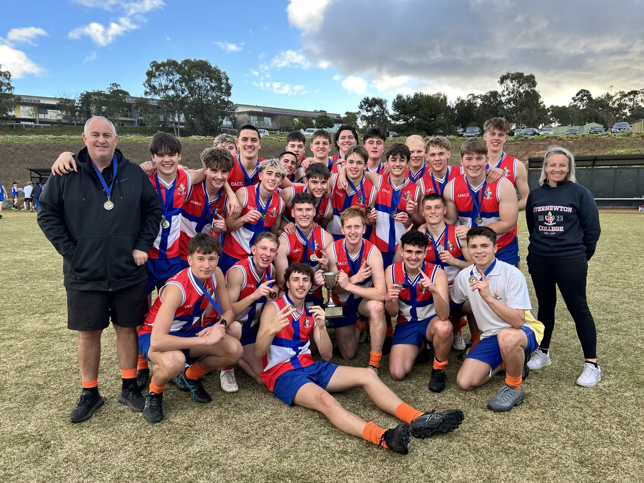 Group shot of the elated Boys 1st Football team flanked by their coaches at the Yirramboi Campus Oval following their 2024 Association of Co-Educational Schools Competition win, proudly holding the winner’s cup.