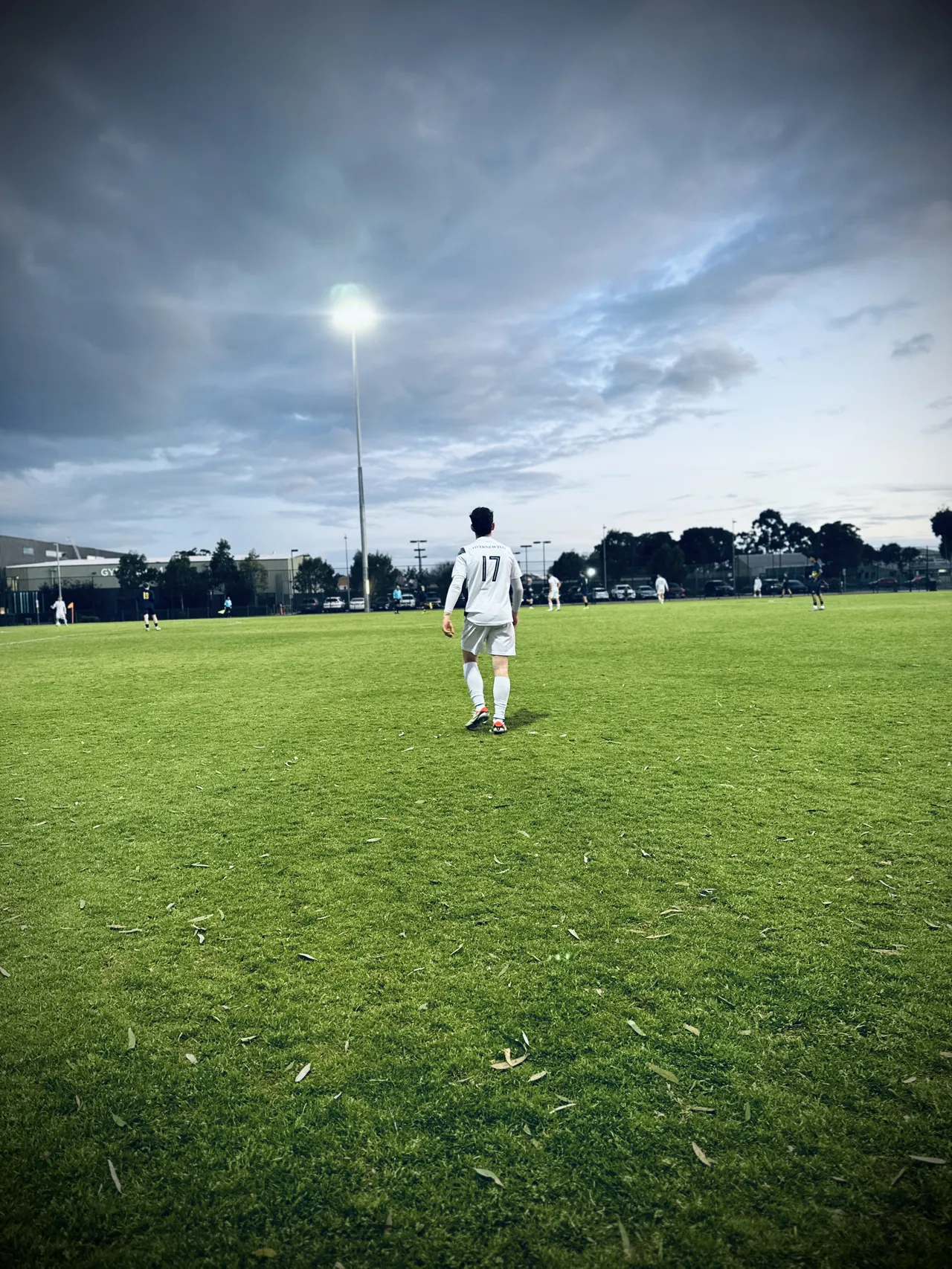 Wide shot of a lone Overnewton upper secondary school aged soccer player walking towards a stormy twilight sky on an empty pitch, in preparation for an Association of Co-Educational Schools night match.