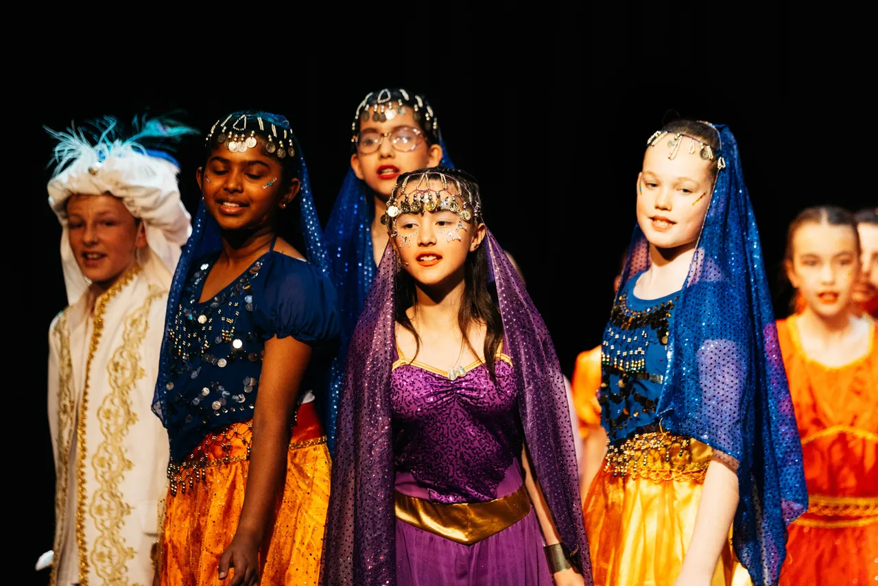 Group of upper primary school aged children in vibrant, opulent costumes performing on stage in the musical Aladdin, as part of the Overnewton's Encore Performing Arts Co Curricular program.