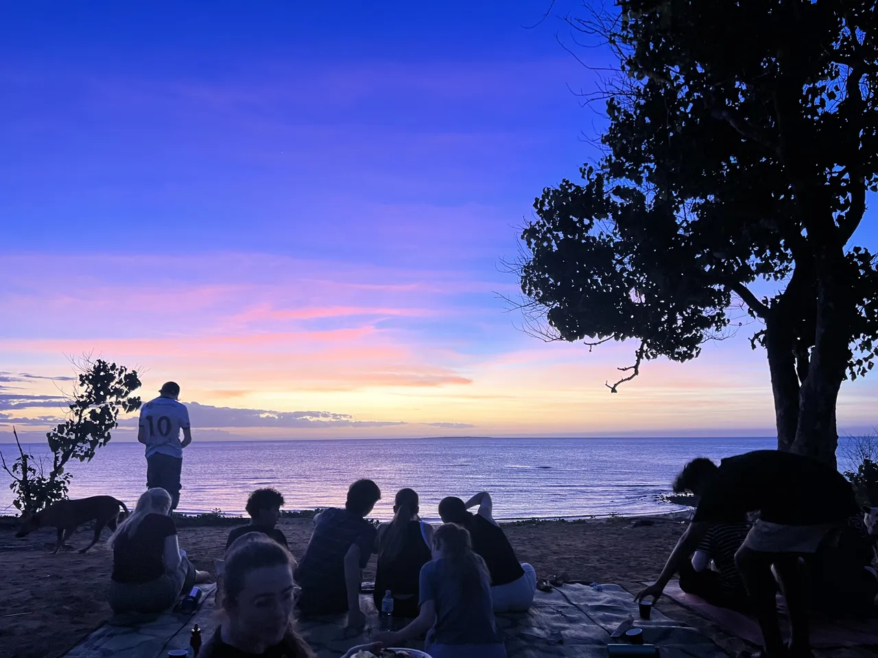 Year 9 and 10 students from Overnewton seated on a picnic rug facing the ocean at sunset, silhouetted against pastel skies during their Northern Territory Learning on Country trip.